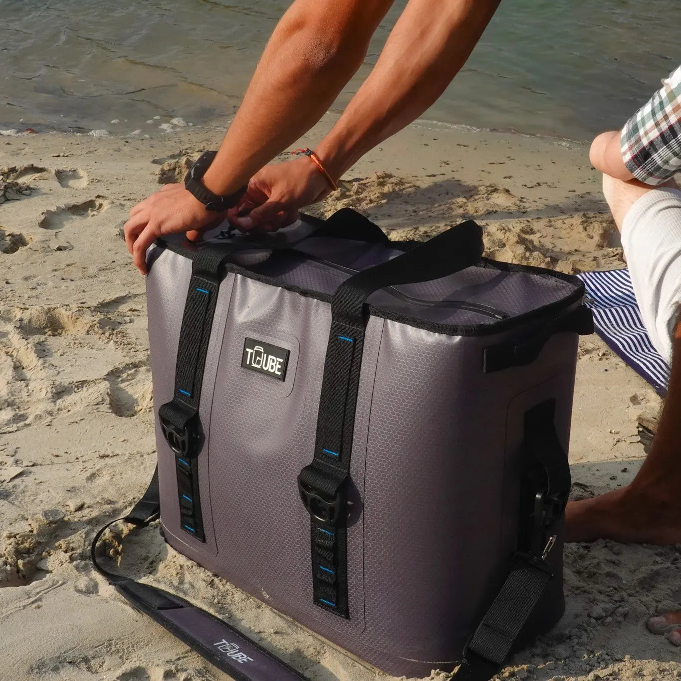 Person holding Tuube Traveller 36L with black straps on a sandy beach, demonstrating shoulder carry comfort for large travel cooler