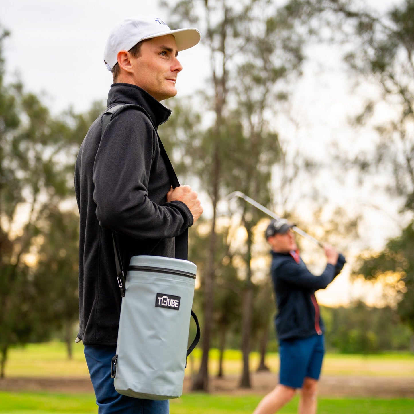 Tuube Cooler Bag XL grey with shoulder strap attached, golfer swinging in background, showing hands-free carry option on the course